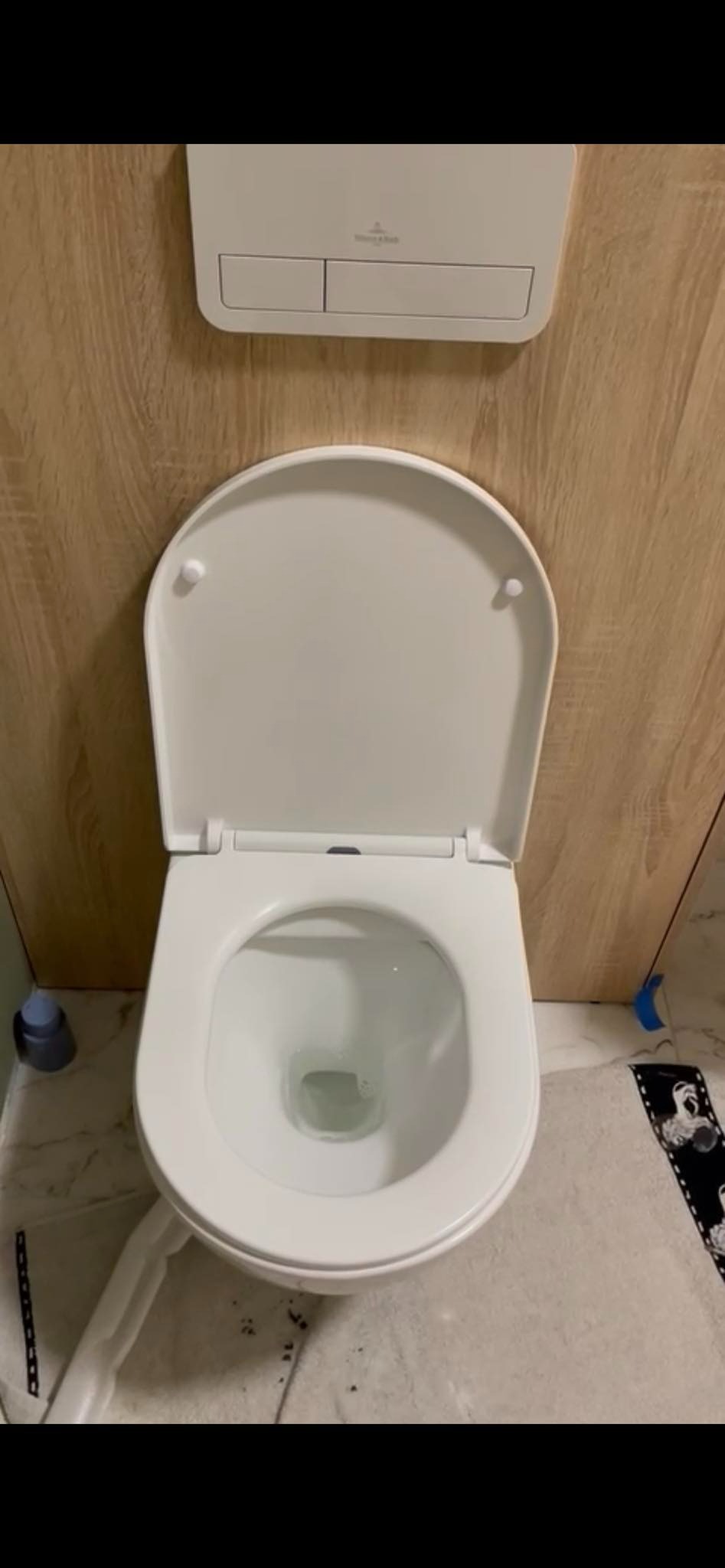 Wisząca toaleta z otwartą deską, widoczny przycisk spłuczki Villeroy & Boch na ścianie z drewnopodobnych paneli, częściowo zakryta podłoga z szarych płytek.