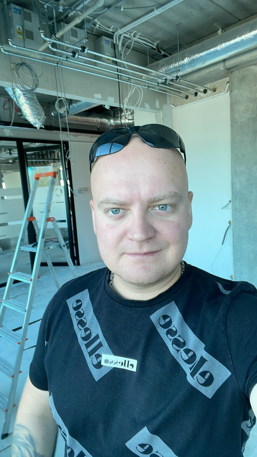 Mężczyzna w okularach przeciwsłonecznych na głowie pozuje na tle instalacji sufitowych i drabiny w trakcie remontu wnętrza.
