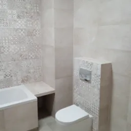 Beżowa łazienka z mozaiką na ścianie, wisząca toaleta i zabudowana wanna z półką, płytki podłogowe w odcieniach szarości.