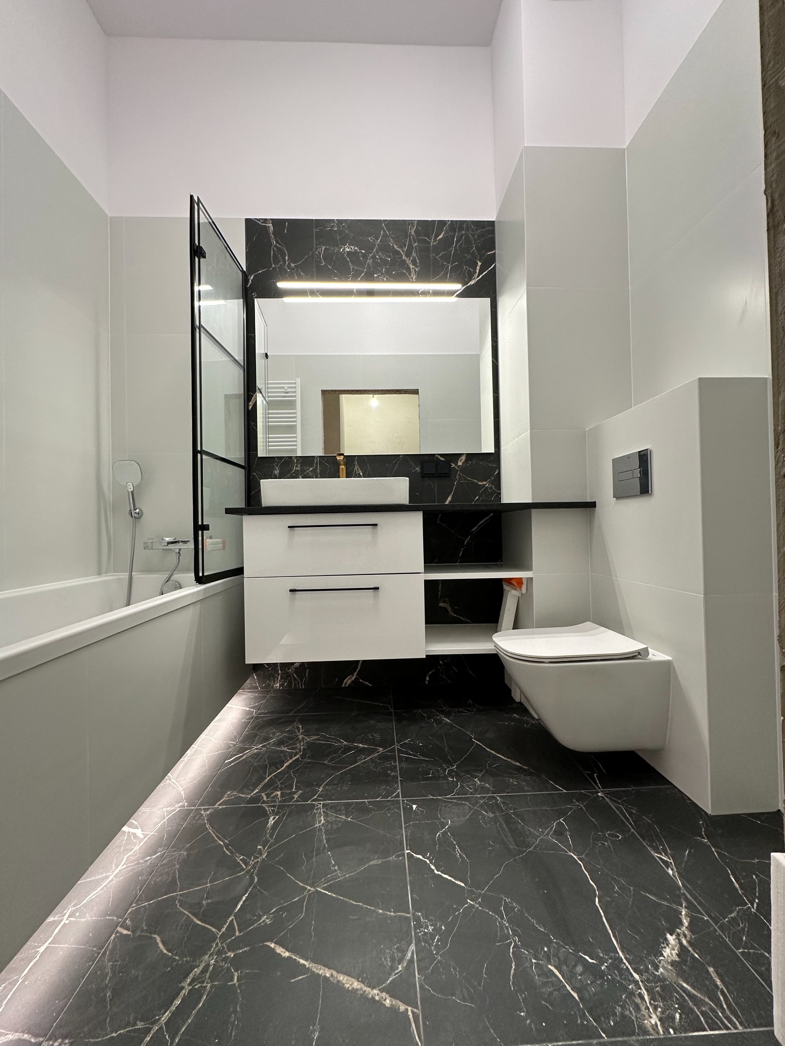 Nowoczesna łazienka z czarno-białymi płytkami imitującymi marmur na podłodze i ścianie za umywalką, białe meble łazienkowe z czarnymi uchwytami, wisząca toaleta i szklany parawan nawannowy...