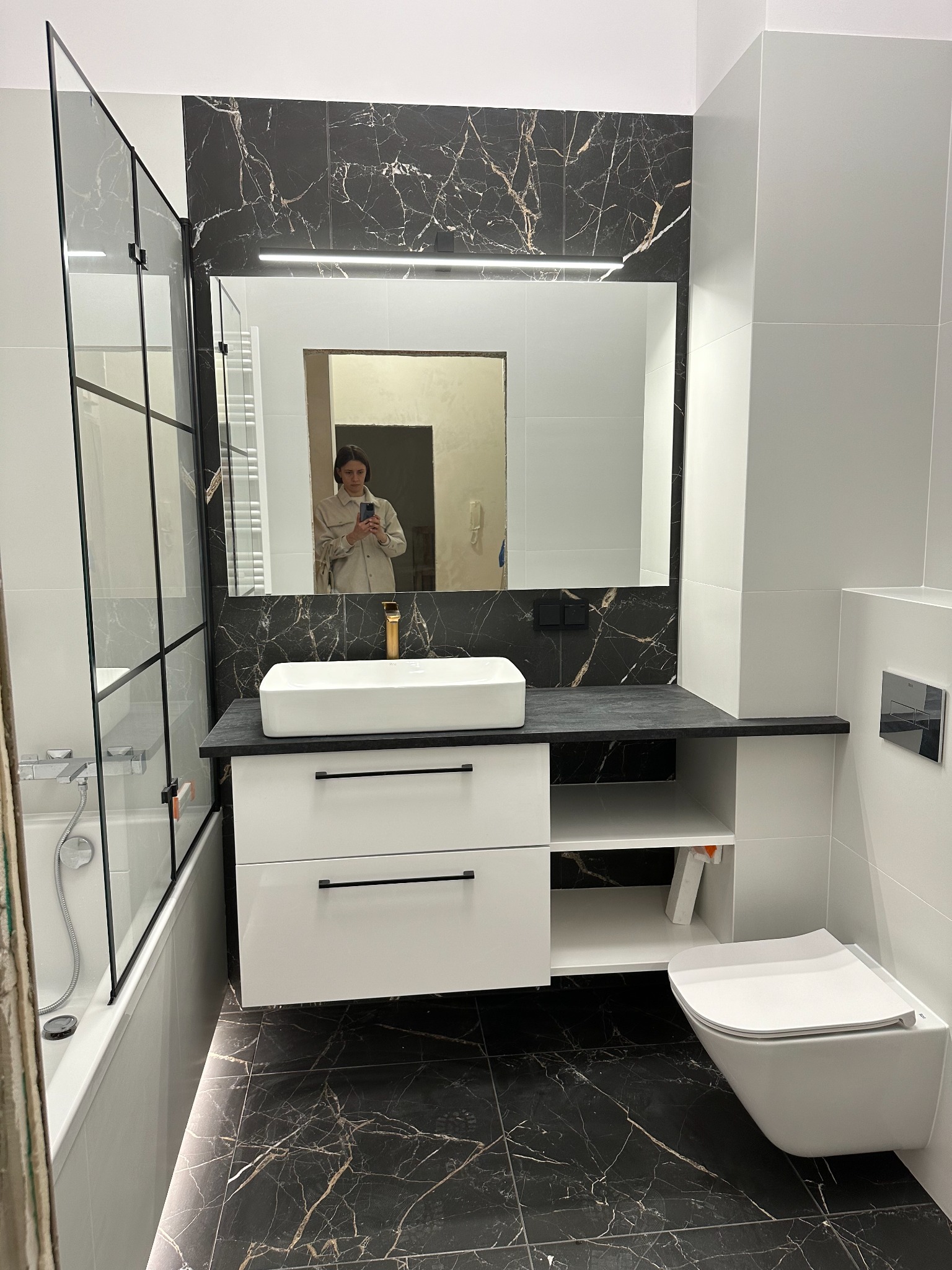 Nowoczesna łazienka z czarno-białymi płytkami imitującymi marmur, kabiną prysznicową ze szklanymi panelami i czarnymi ramami, białą umywalką nablatową na szafce z szufladami i półkami,...