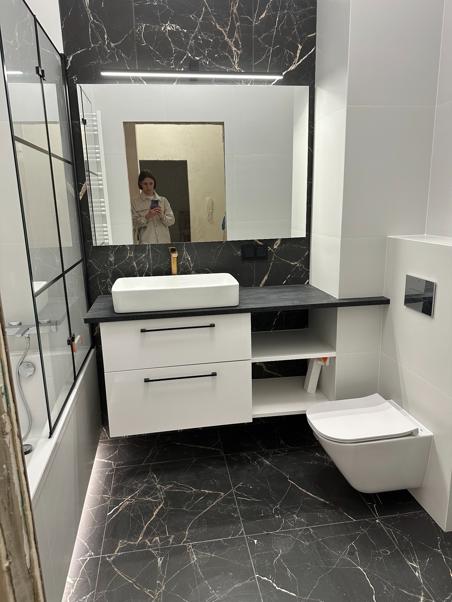 Nowoczesna łazienka z czarno-białymi płytkami imitującymi marmur, białą umywalką nablatową z złotą armaturą, szafką z szufladami i wiszącą toaletą; w lustrze odbija się osoba robiąca zdjęcie.