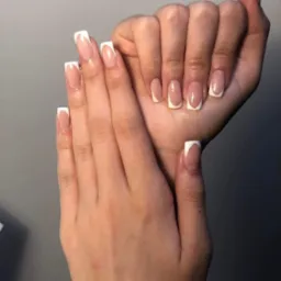 Dłonie prezentują świeżo wykonany manicure francuski z kwadratowymi końcówkami, subtelny połysk na paznokciach, neutralne tło.