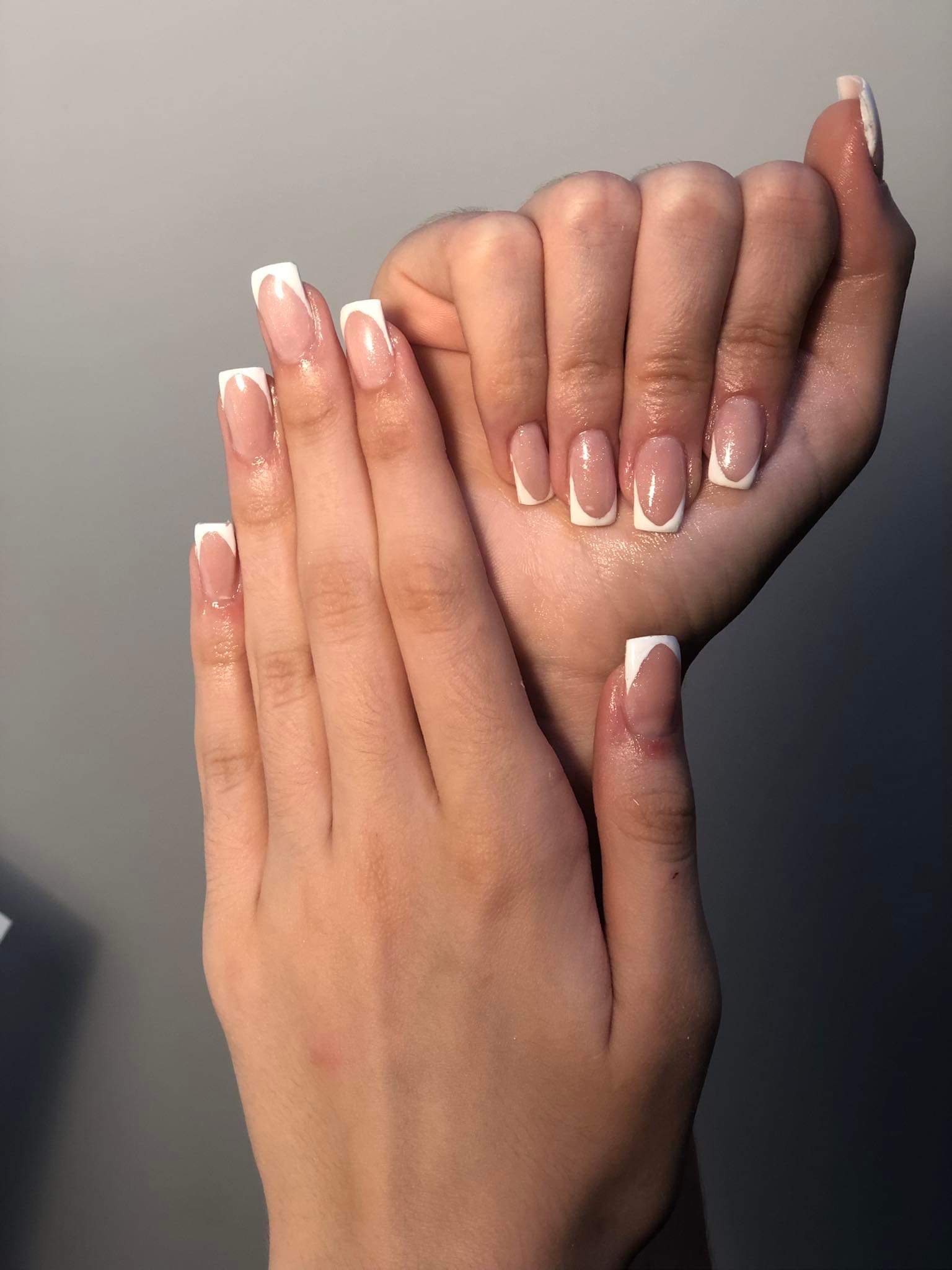 Dłonie prezentują świeżo wykonany manicure francuski z kwadratowymi końcówkami, subtelny połysk na paznokciach, neutralne tło.