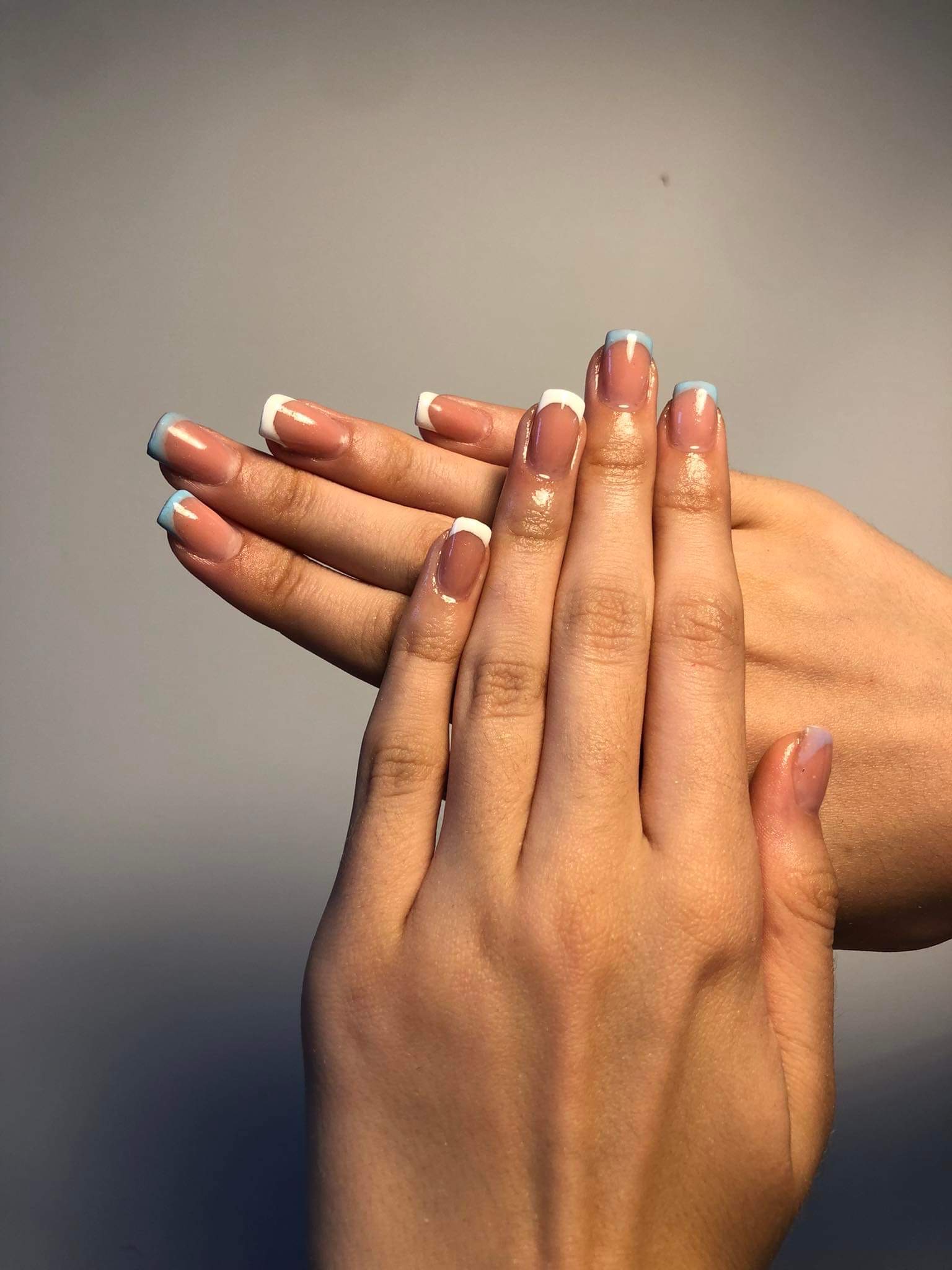 Dłonie z kwadratowymi paznokciami w stylu french manicure: białe końcówki z dodatkiem błękitnego akcentu, widoczne delikatne refleksy światła na lakierze.