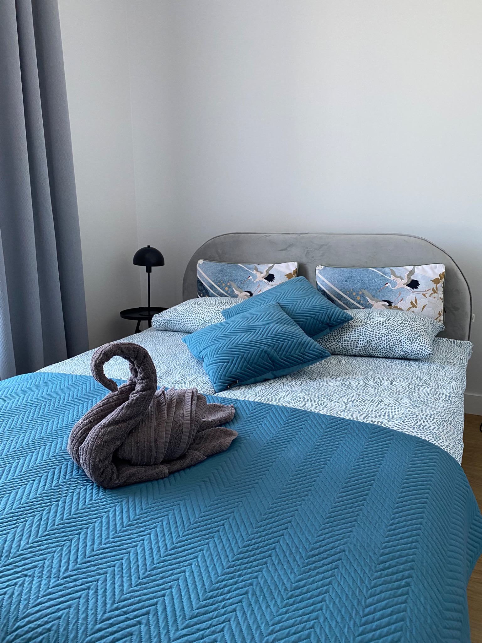Ustawione łóżko w pokoju hotelowym z ręcznikiem uformowanym w łabędzia, błękitną narzutą w jodełkę i poduszkami z motywem żurawi. Minimalistyczny wystrój.