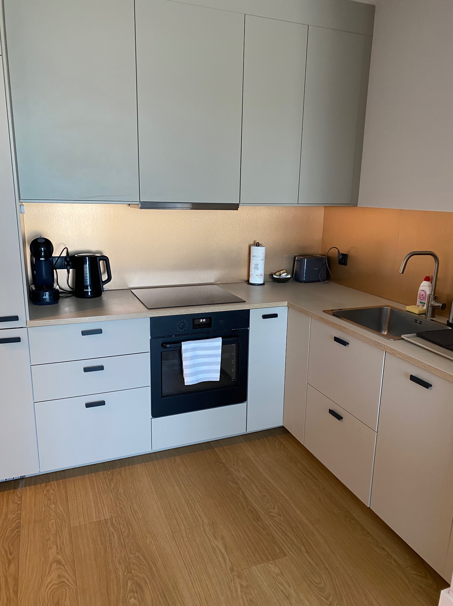 Nowoczesna, jasna kuchnia z drewnianą podłogą. Zabudowa kuchenna w odcieniach beżu i szarości, z czarnymi uchwytami. Na blacie ekspres do kawy i czajnik.