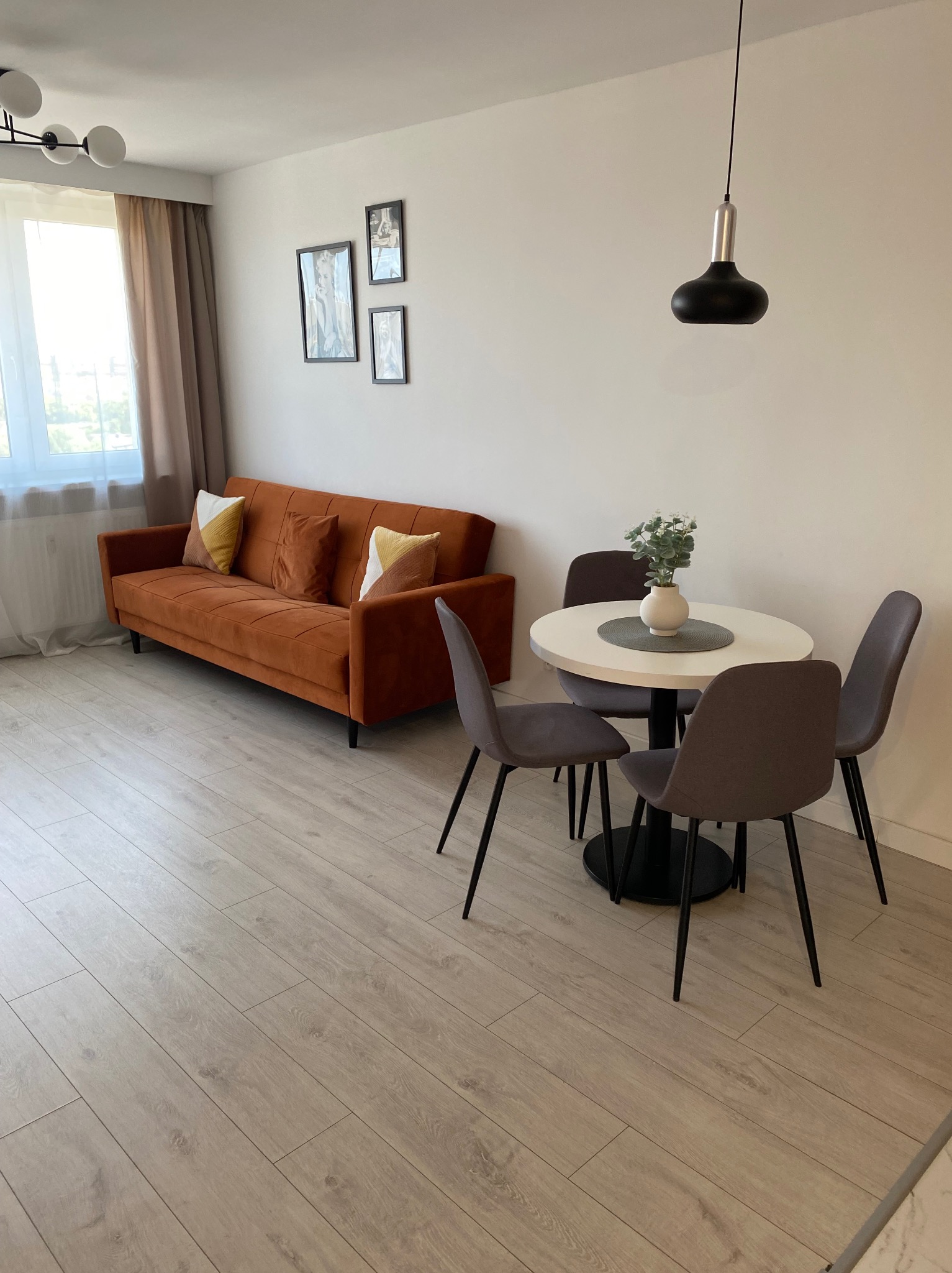 Jasne, nowoczesne wnętrze: pomarańczowa sofa z poduszkami, okrągły stół z kwiatami i szare krzesła na drewnianej podłodze, doświetlone światłem z okna.