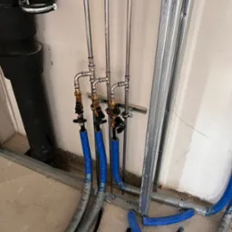 Szare i niebieskie rury instalacyjne z zaworami, prowadzone wzdłuż ściany z metalowym stelażem, widoczne elementy mocujące i fragment podłogi w trakcie prac instalacyjnych.