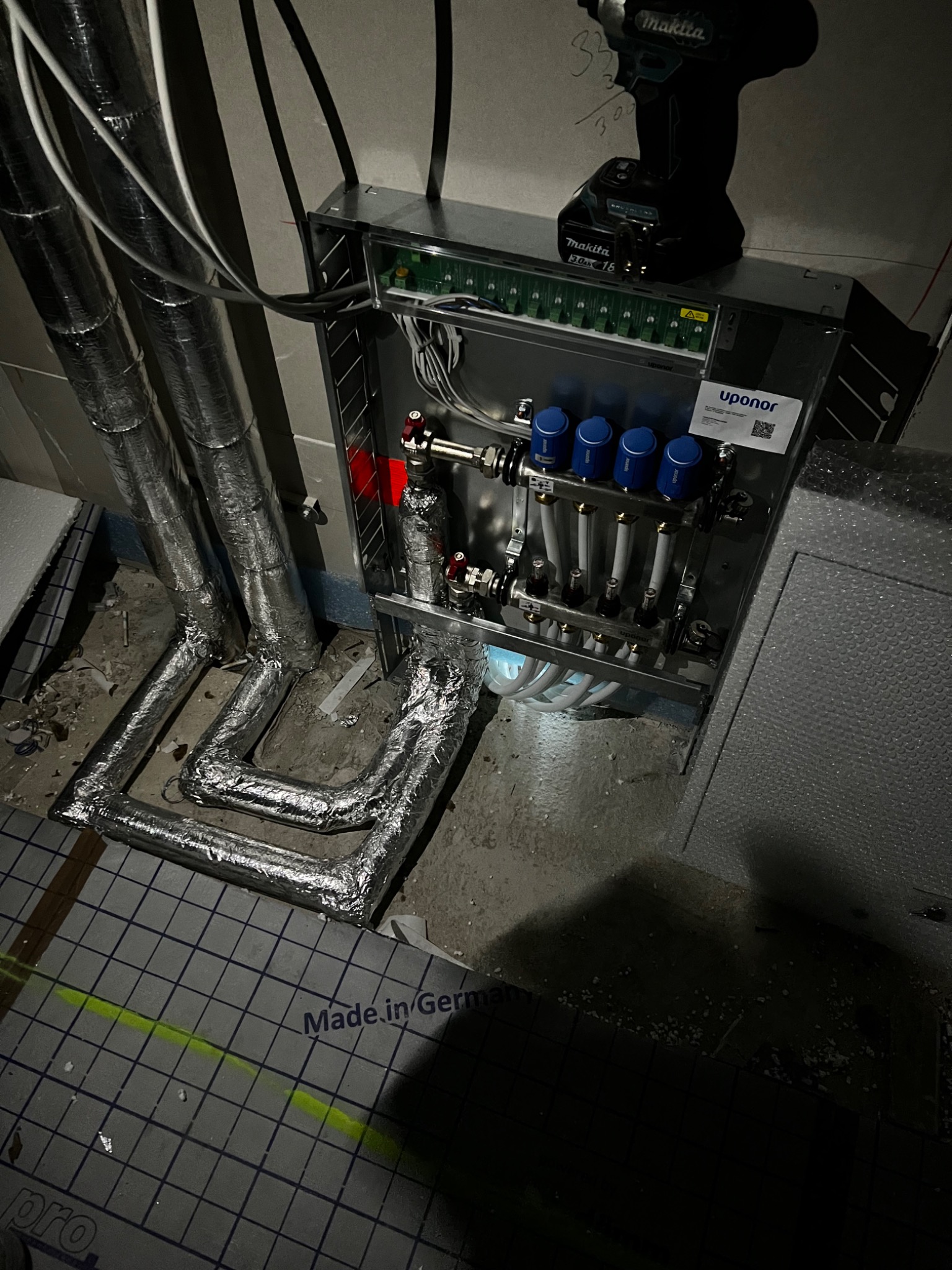 Rozdzielacz ogrzewania podłogowego Uponor w metalowej szafce, z niebieskimi zaworami i białymi rurkami, podłączony do izolowanych rur aluminiowych, z wkrętarką Makita na górze i napisem 'Made in...