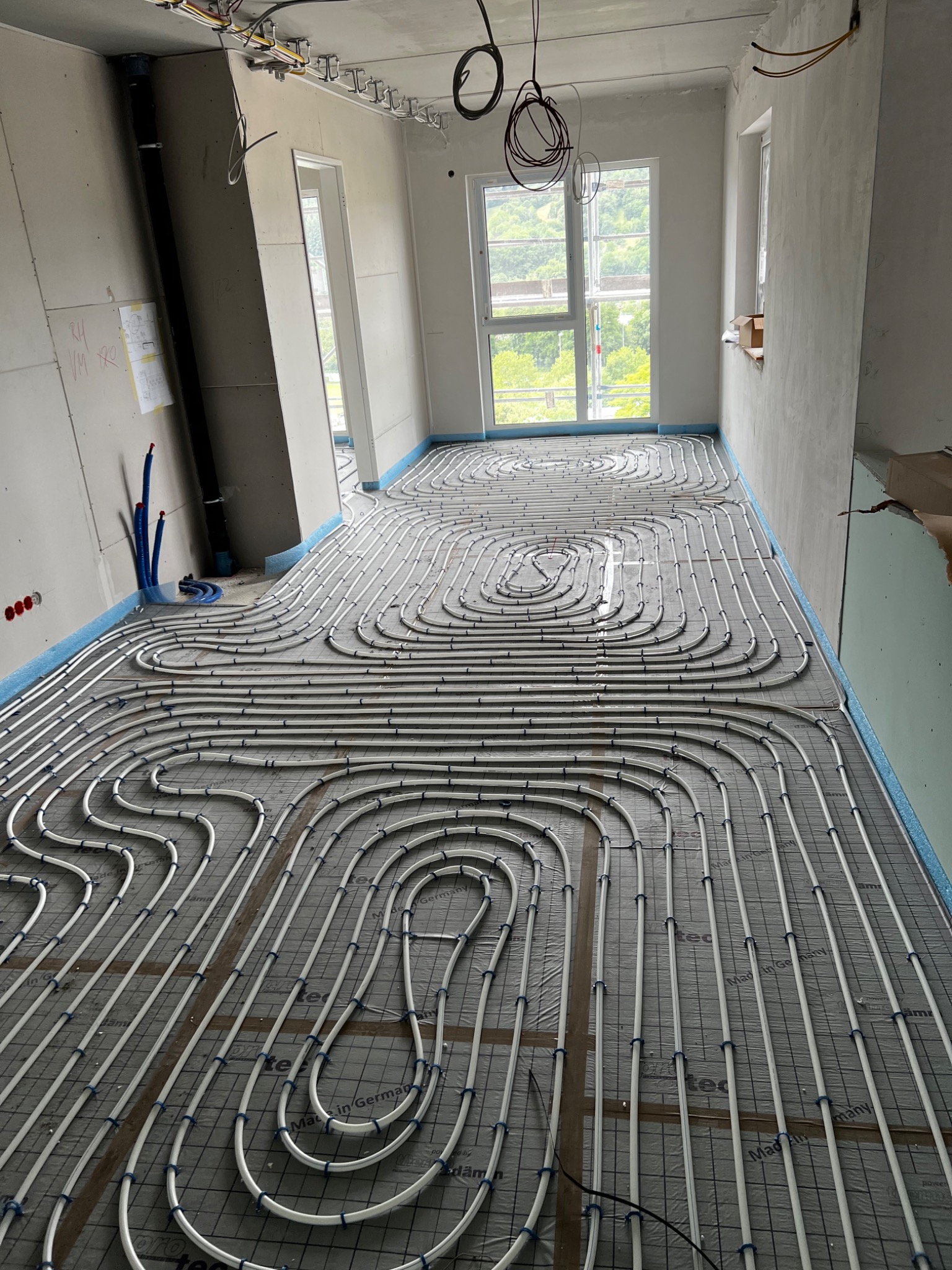 Rozłożone białe rurki systemu grzewczego podłogowego na szarej macie z niebieskimi brzegami w surowym wnętrzu podczas budowy, widoczne okna i wystające kable.