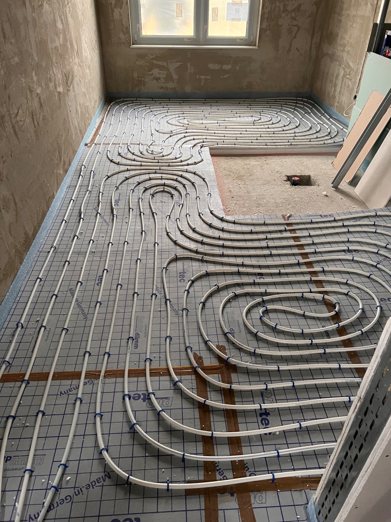 Rozłożone białe rury ogrzewania podłogowego na izolacyjnej macie z niebieską siatką, widoczne mocowania rur, surowe ściany w tle, okno z widoczną folią ochronną, fragment niezabezpieczonej podłogi...
