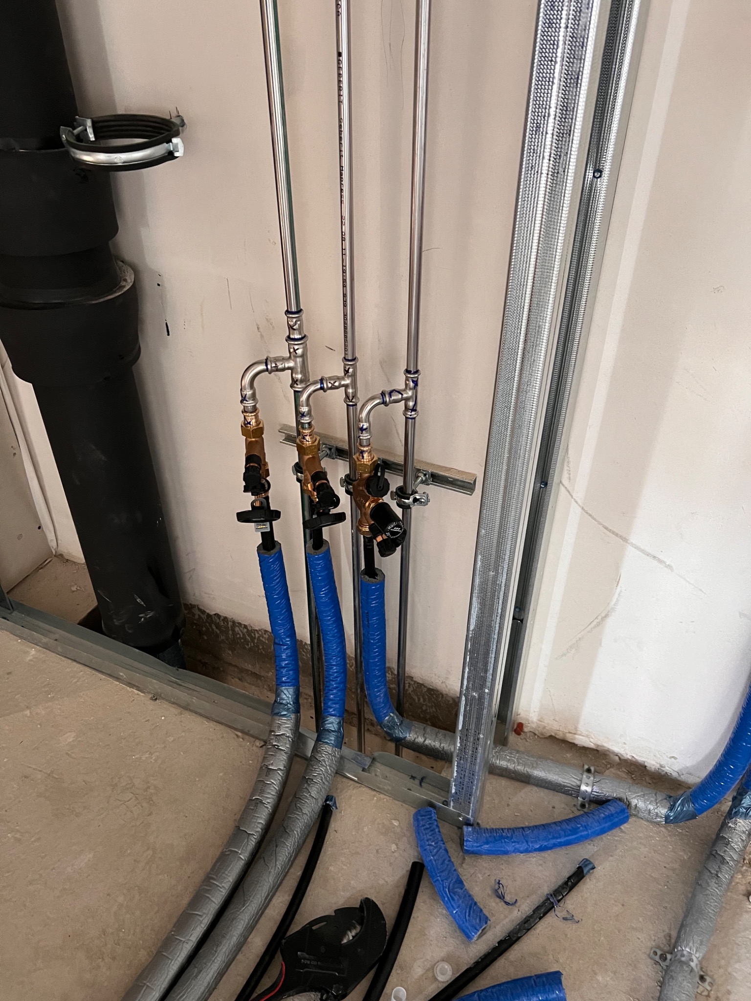 Szare i niebieskie rury instalacyjne z zaworami, prowadzone wzdłuż ściany z metalowym stelażem, widoczne elementy mocujące i fragment podłogi w trakcie prac instalacyjnych.
