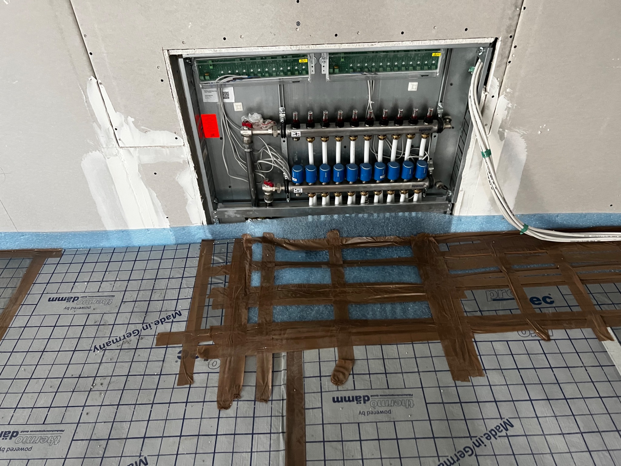 Rozdzielacz systemu ogrzewania podłogowego Uponor zamontowany w ścianie, widoczne rury i zawory, poniżej izolacja pod wylewkę z folią rastrową i taśmą mocującą. Na folii widoczny napis Made in...