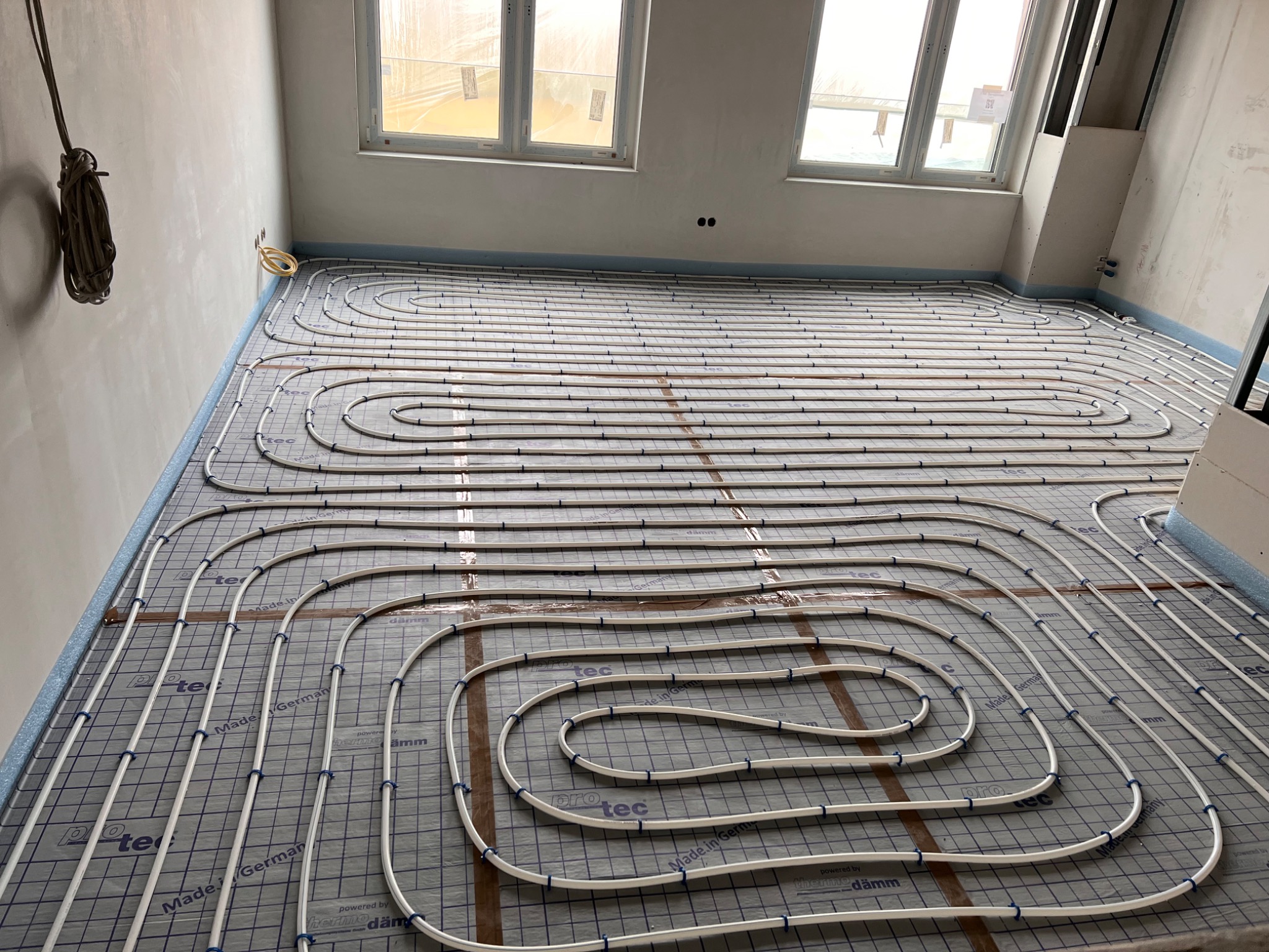 Białe rurki systemu ogrzewania podłogowego rozłożone spiralnie na izolacji podłogowej z nadrukiem siatki, widoczne okna i surowe ściany w tle, przygotowanie do wylewki.