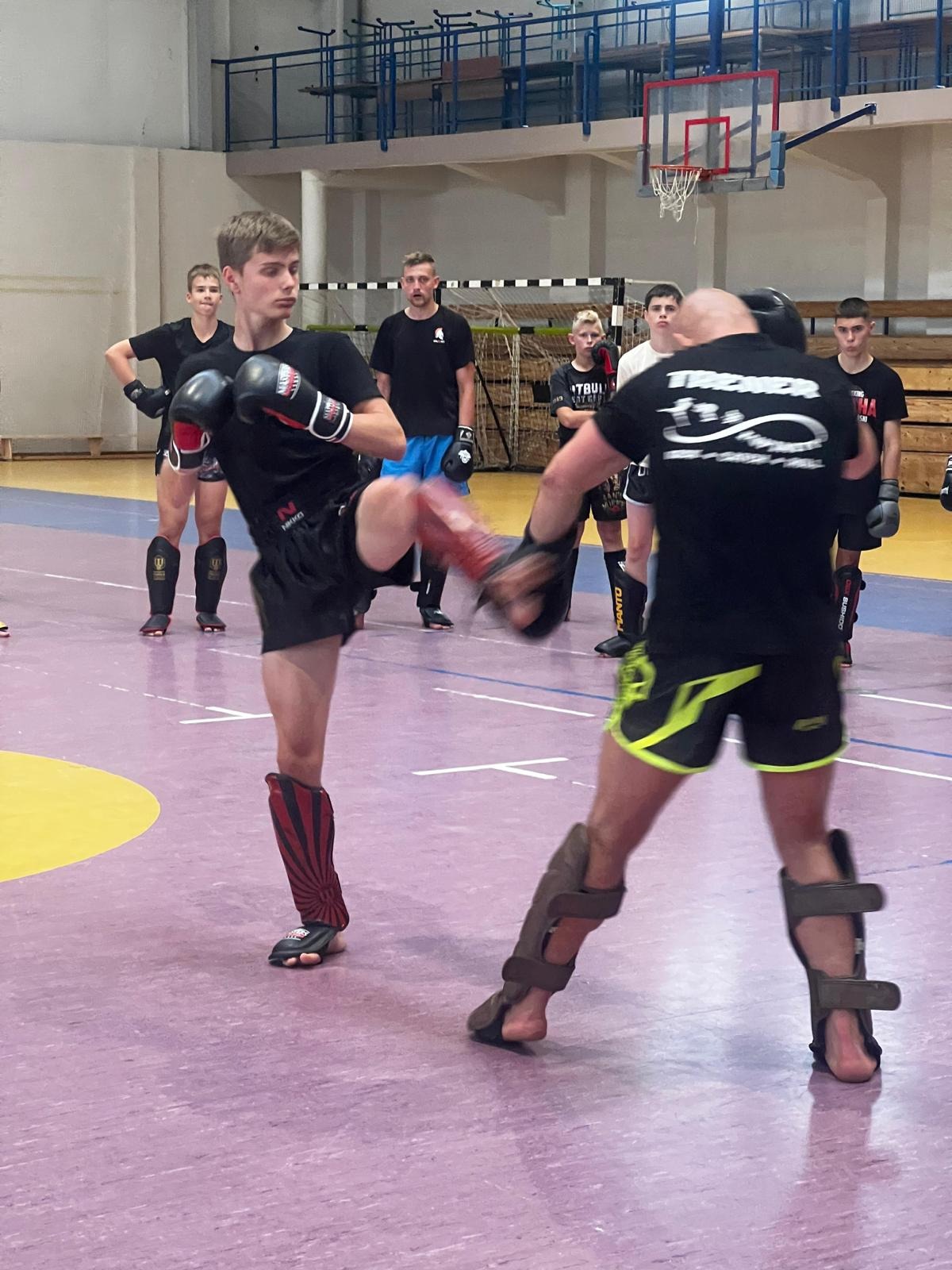 Młody zawodnik w czarnych spodenkach i rękawicach, z czerwoną ochraniaczem na nodze, wykonuje kopnięcie w kierunku trenera z tarczą, podczas treningu na sali gimnastycznej, w tle grupa obserwujących.