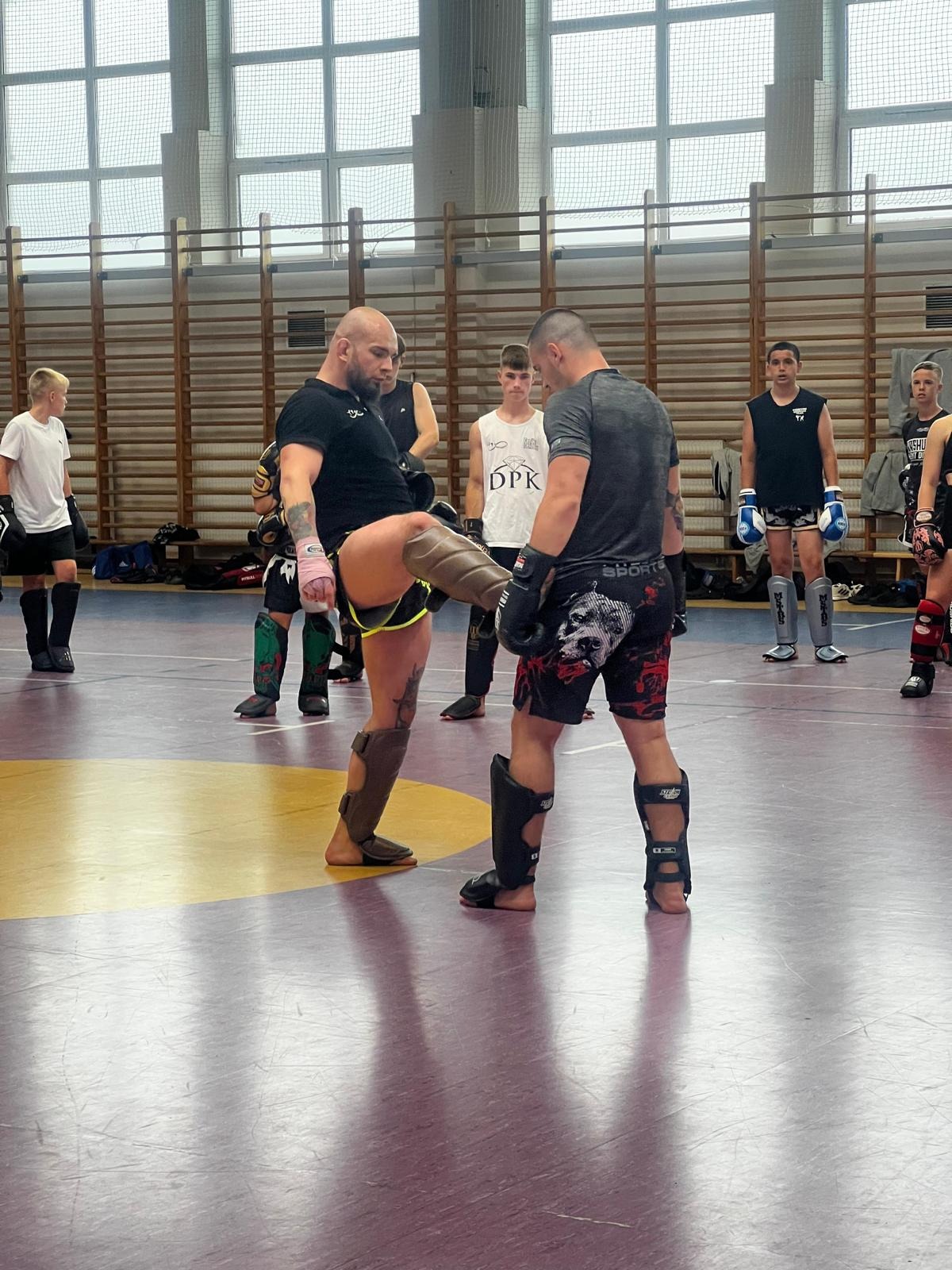 Dwóch mężczyzn w ochraniaczach na nogi i rękawicach bokserskich w trakcie sparingu na sali gimnastycznej, w tle grupa obserwujących.