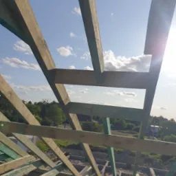 Drewniana więźba dachowa z widocznym niebem i chmurami w tle, świeżo zaimpregnowana na zielono