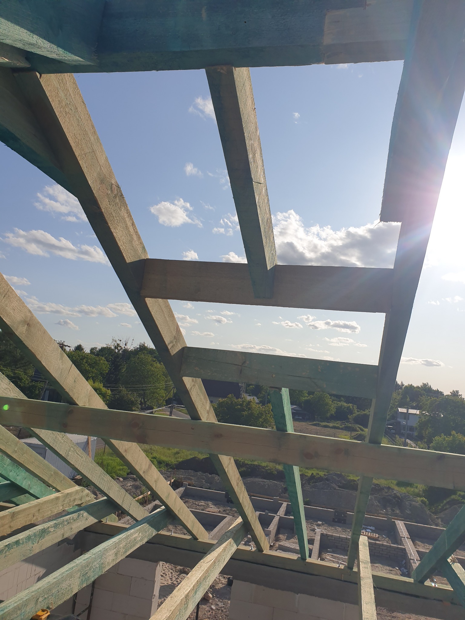 Drewniana więźba dachowa z widocznym niebem i chmurami w tle, świeżo zaimpregnowana na zielono