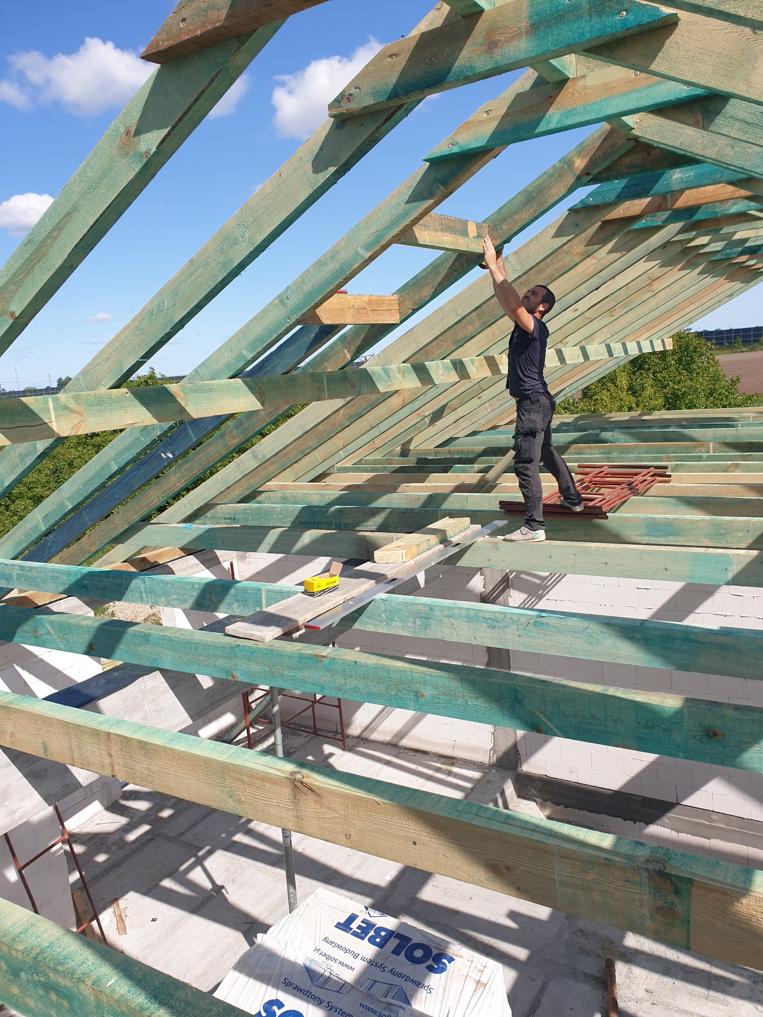 Pracownik montuje elementy drewnianej konstrukcji dachu z zielonym impregnatem, widoczne narzędzia i częściowo ułożona dachówka, błękitne niebo w tle.