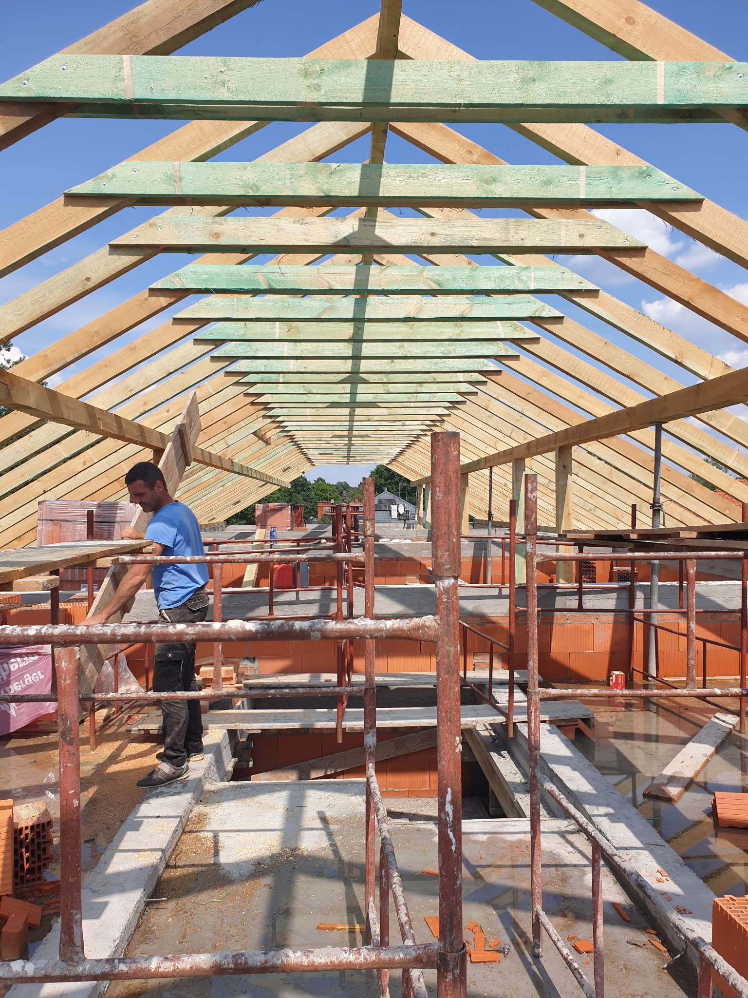 Montaż drewnianej konstrukcji dachu z widocznymi elementami impregnowanymi na zielono, pracownik w niebieskiej koszulce przenosi deskę na tle błękitnego nieba.