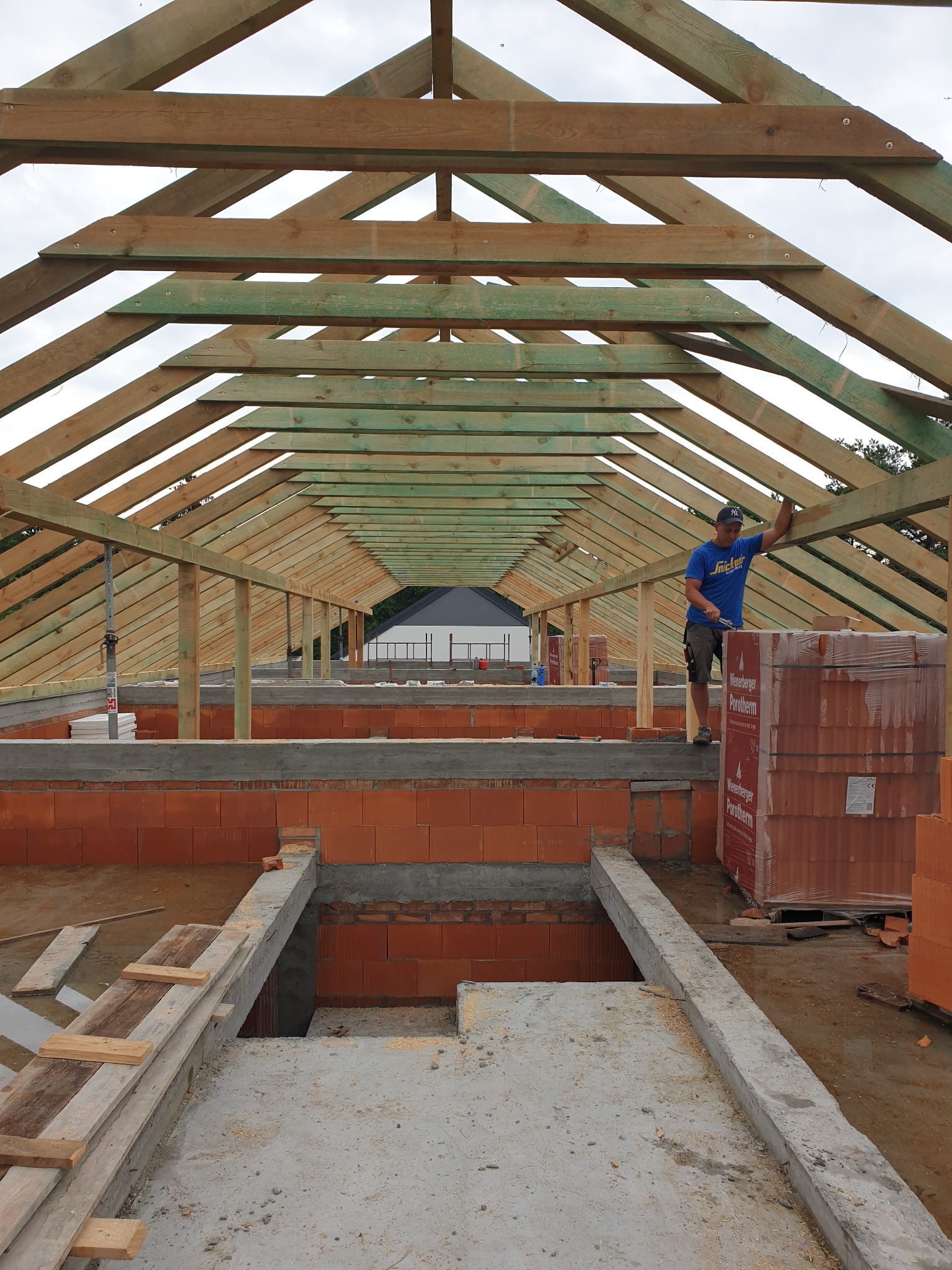 Konstrukcja drewniana więźby dachowej w trakcie montażu w Milanówku, widok z wnętrza budynku, pracownik w niebieskiej koszulce na tle belek i cegieł Porotherm.