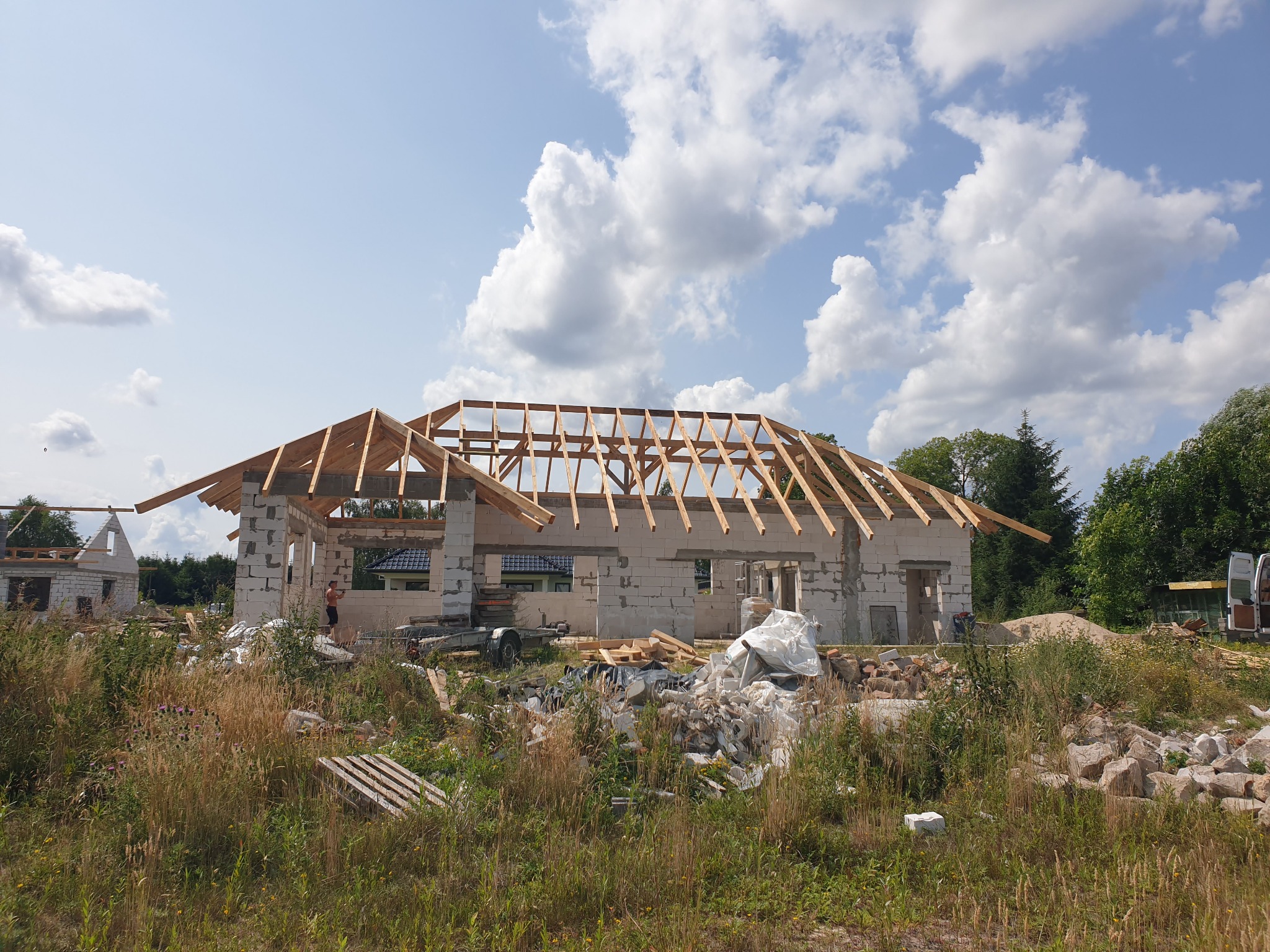 Wznoszenie więźby dachowej na surowym budynku z białego betonu komórkowego, widok z zarośniętego placu budowy w słoneczny dzień.