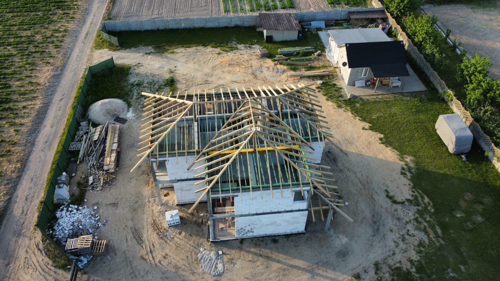 Wysoka perspektywa budowy domu jednorodzinnego: surowy stan z widoczną konstrukcją drewnianą więźby dachowej, białe bloczki ścian, materiały budowlane i otoczenie działki.