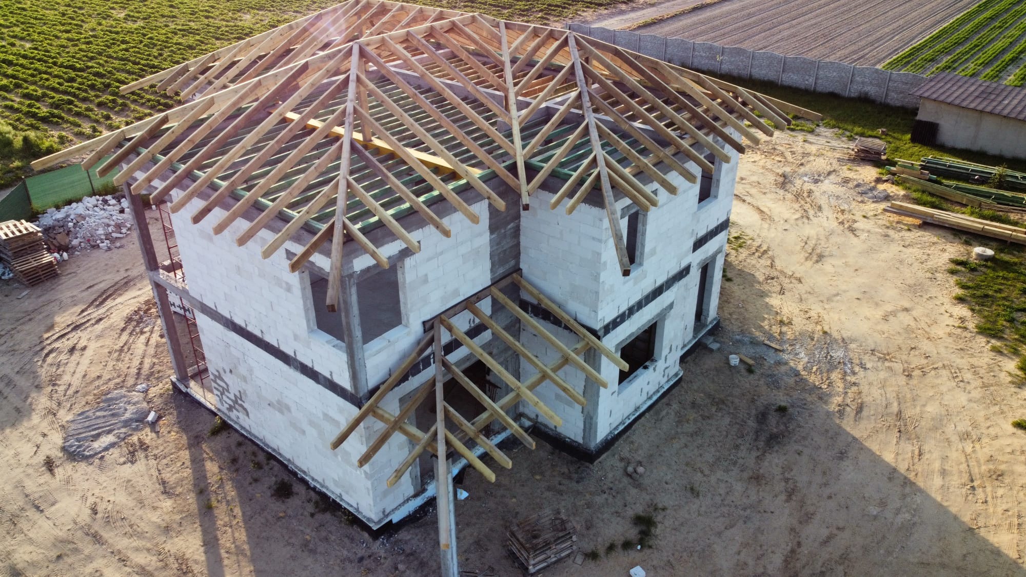 Surowy stan domu w trakcie budowy, widok z góry na konstrukcję drewnianą dachu z elementami więźby dachowej, ściany z pustaków, palety na ziemi.