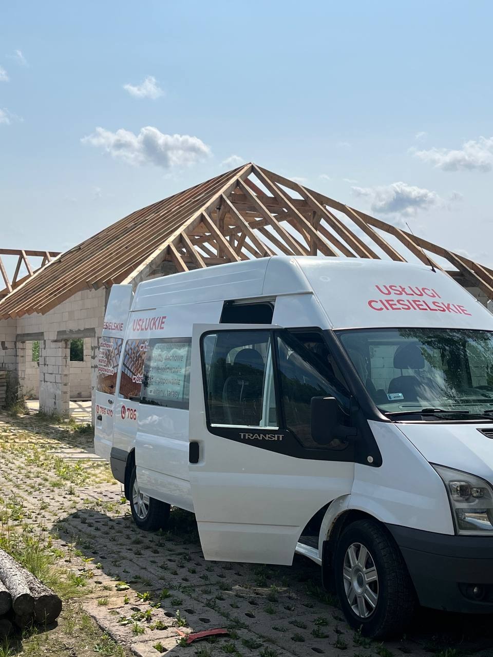 Wznoszona więźba dachowa na budynku w trakcie budowy, widoczny biały bus z reklamą usług ciesielskich i otwarte drzwi, na pierwszym planie stos drewna.