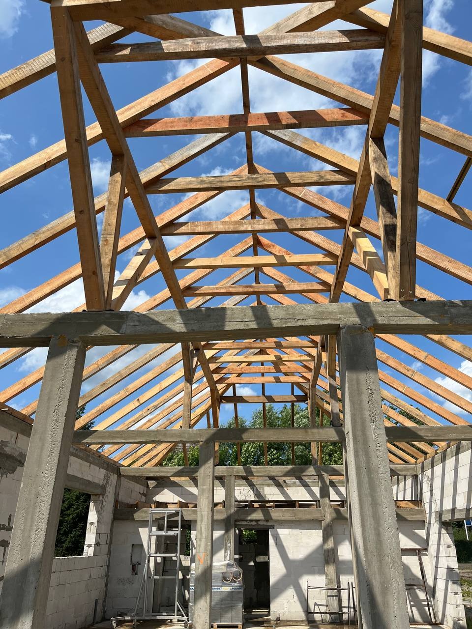 Konstrukcja drewnianej więźby dachowej w trakcie montażu, widok od wewnątrz budynku z betonu komórkowego, błękitne niebo w tle.