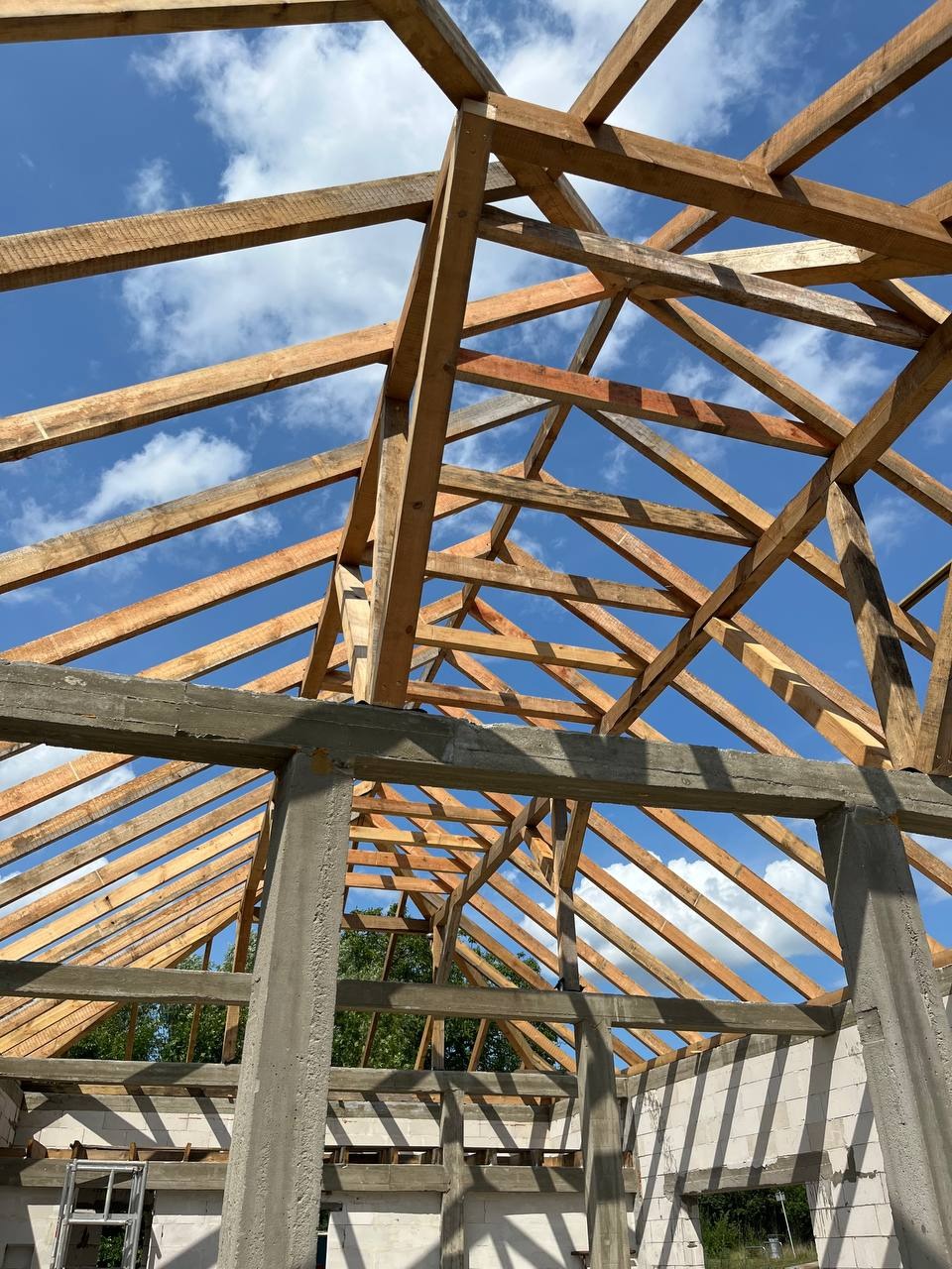 Surowa konstrukcja drewnianej więźby dachowej na tle błękitnego nieba z chmurami, widok od dołu, częściowo z betonu i bloczków, budynek w stanie surowym otwartym.