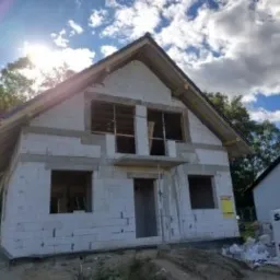 Surowy stan domu z białych bloczk&oacute;w, widoczne niezamontowane okna, drewniana konstrukcja dachu i paleta bloczk&oacute;w Solbet na pierwszym planie, w tle zieleń i błękitne niebo z chmurami.