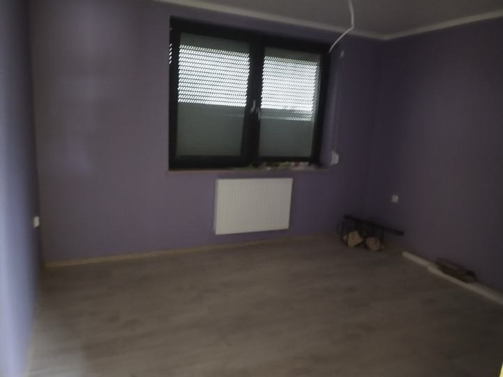 Pusty pokój z fioletowymi ścianami, drewnopodobną podłogą i grzejnikiem pod oknem z żaluzjami. W rogu leżą narzędzia.