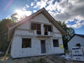 Surowy stan domu z białych bloczków, widoczne niezamontowane okna, drewniana konstrukcja dachu i paleta bloczków Solbet na pierwszym planie, w tle zieleń i błękitne niebo z chmurami.