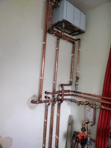 Instalacja grzewcza z miedzianych rur, widoczny bojler oraz pompa obiegowa, ściana w trakcie prac wykończeniowych.