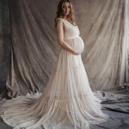 Ewa Krewska Fotografia - Kobieta w zwiewnej, kremowej sukni ciążowej pozuje na tle płóciennej draperii w odcieniach beżu i szarości, trzymając dłonie na brzuchu.
