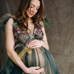 Ewa Krewska Fotografia - Kobieta w kwiecistej sukni z tiulu, trzymająca dłonie na ciążowym brzuchu, spoglądająca w dół, na tle teksturowanej szarej tkaniny.