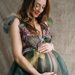 Ewa Krewska Fotografia - Kobieta w ciąży w tiulowej sukni z haftowanymi kwiatami i piórami, dłońmi obejmuje brzuch, spoglądając w dół, na tle beżowej tkaniny.