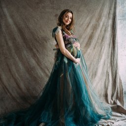 Ewa Krewska Fotografia - Kobieta w ciąży w długiej, tiulowej sukni z kwiatowymi aplikacjami, pozująca na tle teksturowanej tkaniny i ściany.
