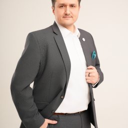 Ewa Krewska Fotografia - Mężczyzna w szarym garniturze, białej koszuli i z turkusową poszetką, pozuje z ręką w kieszeni, prezentując profesjonalny wizerunek na jasnym tle.