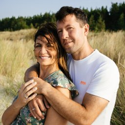 Ewa Krewska Fotografia - Uśmiechnięta para na tle wydm i lasu. Kobieta w kwiecistej sukience, mężczyzna w białym t-shircie obejmuje ją. Widoczne obrączki. Naturalne światło, ciepła kolorystyka.