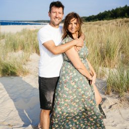 Ewa Krewska Fotografia - Uśmiechnięta para na plaży w słoneczny dzień. Mężczyzna obejmuje kobietę w kwiecistej sukience. W tle wydmy i morze.