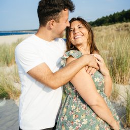 Ewa Krewska Fotografia - Uśmiechnięta para na plaży, mężczyzna obejmuje kobietę w kwiecistej sukience, w tle wydmy i morze pod błękitnym niebem. Romantyczna sesja w naturalnym świetle.