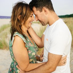 Ewa Krewska Fotografia - Uśmiechnięta para, kobieta w kwiecistej sukience i mężczyzna w białym t-shircie, przytuleni na tle plaży z trawami i morzem w oddali, delikatne światło.