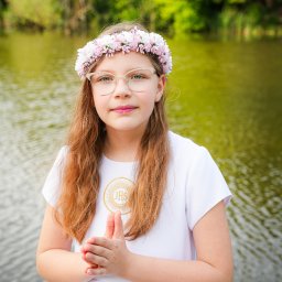 Ewa Krewska Fotografia - Dziewczynka w okularach i wianku z różowych kwiatów, ubrana w białą sukienkę komunijną z symbolem JHS, na tle zieleni i wody. Ujęcie portretowe, skupione na twarzy.
