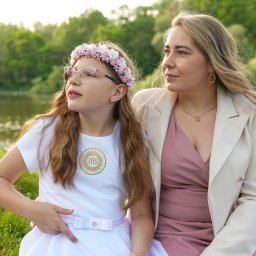 Ewa Krewska Fotografia - Dziewczynka w okularach i wianku, ubrana w białą sukienkę komunijną, siedzi obok kobiety w różowej sukience i beżowej marynarce na tle jeziora i zieleni.