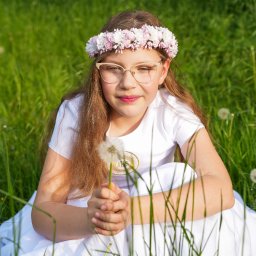 Ewa Krewska Fotografia - Dziewczynka w okularach i wianku na łące, ubrana w białą sukienkę i koszulkę z symbolem IHS, trzyma dmuchawca. Portret w naturalnym świetle.