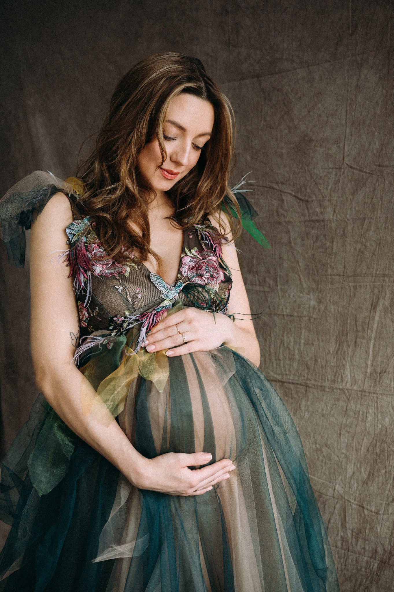 Kobieta w kwiecistej sukni z tiulu, trzymająca dłonie na ciążowym brzuchu, spoglądająca w dół, na tle teksturowanej szarej tkaniny.