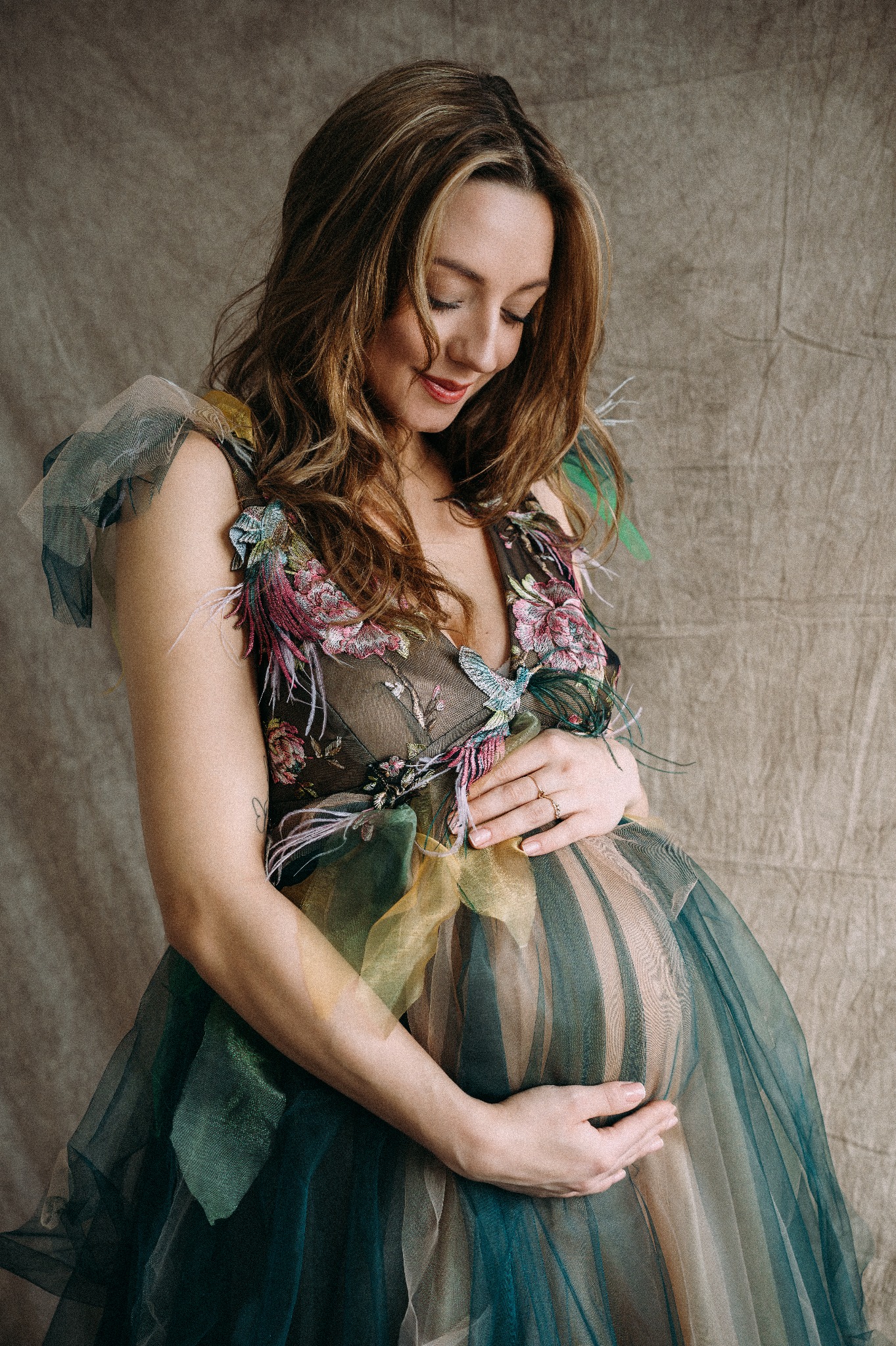 Kobieta w ciąży w tiulowej sukni z haftowanymi kwiatami i piórami, dłońmi obejmuje brzuch, spoglądając w dół, na tle beżowej tkaniny.